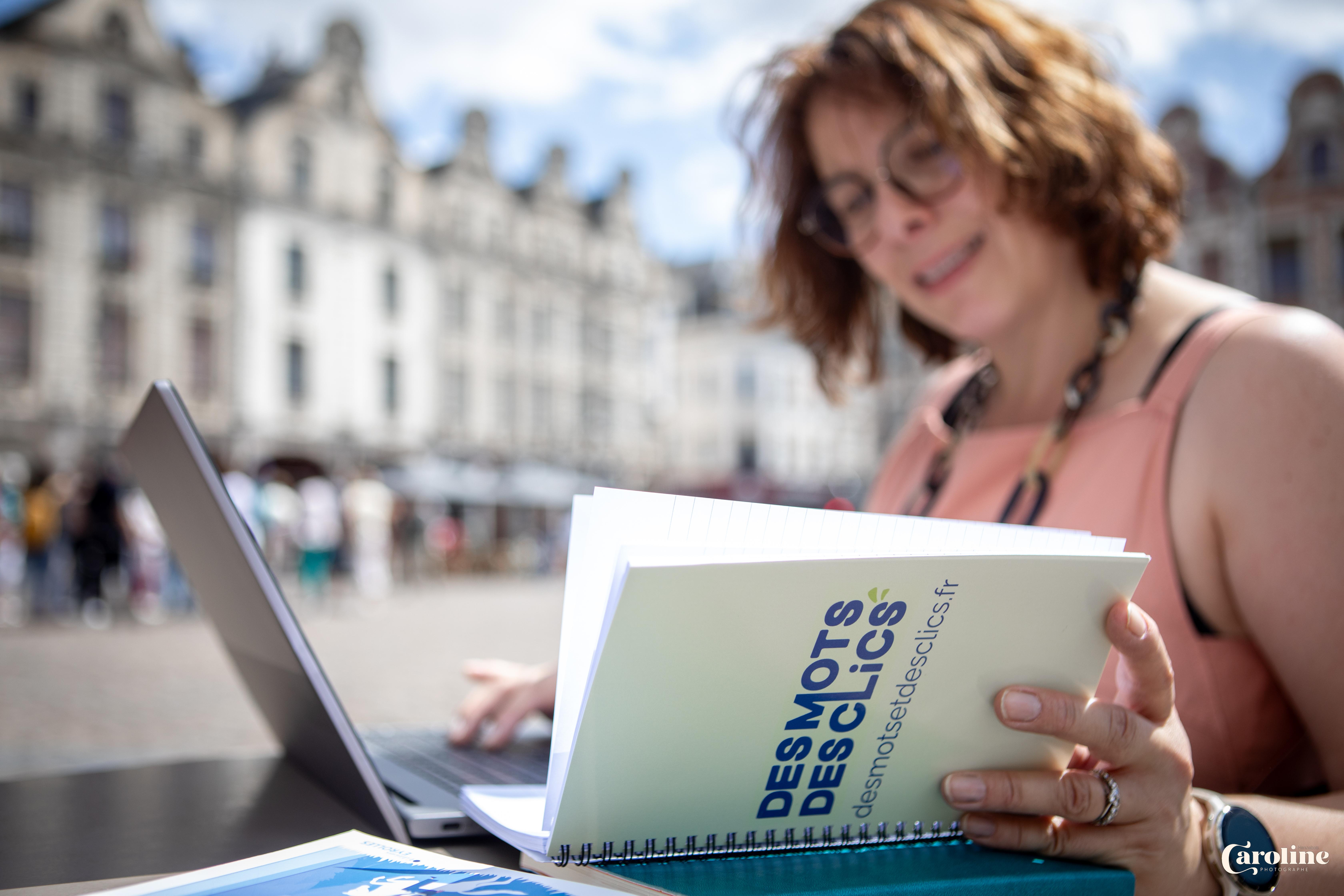 Photo de Gaëlle, la consultante digitale. elle est en train de travailler en extérieur . elle a son ordinateur devant elle et consulte son carnet sur lequel est noté le nom de sa société : Des Mots et des Clics. Elle prépare une stratégie éditoriale pour l'un de ses clients.